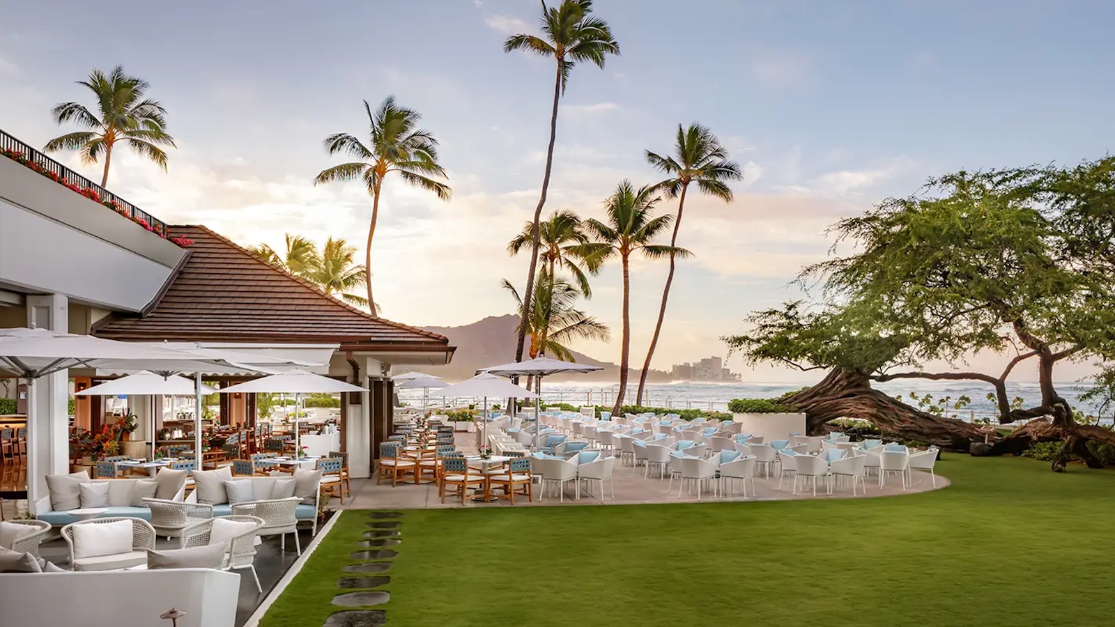 Outdoor dining area at a beachside restaurant with white tables and chairs, surrounded by lush green grass and palm trees. In the background, there's a view of the ocean and distant mountains under a clear sky during sunset—reminiscent of the best hotels with pools in Hawaii.