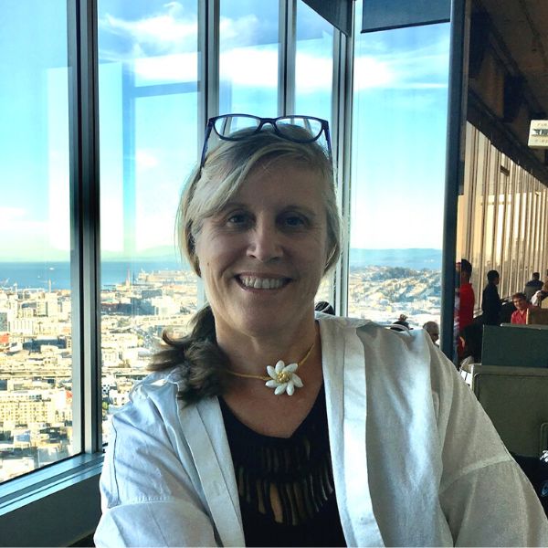 A smiling woman with glasses resting on her head sits by a large window with a cityscape view behind her. She is wearing a light-colored jacket and a floral necklace. Other people are visible in the background enjoying the view.