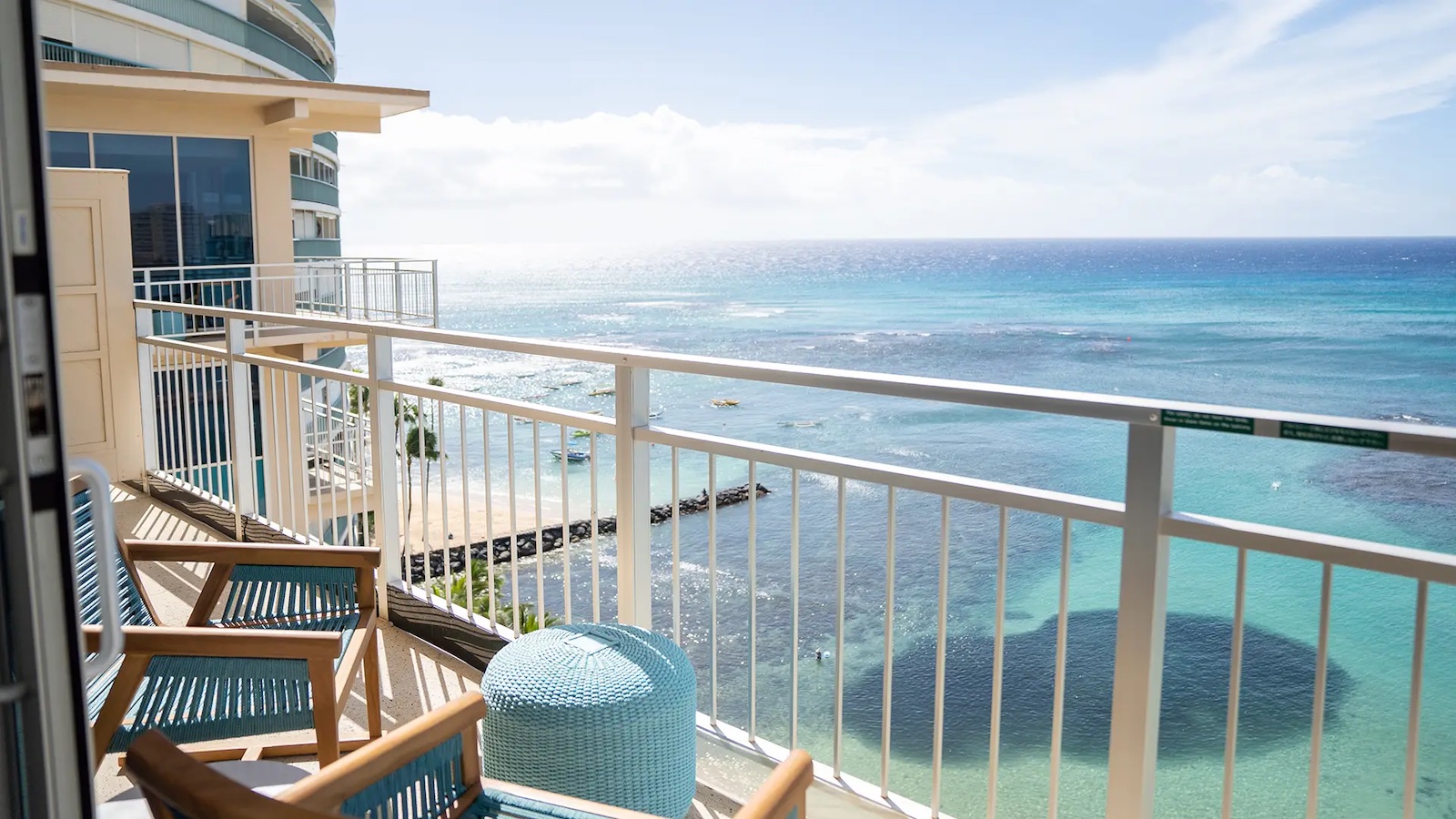 Balcony view from one of the value hotels in the Hawaiian Islands, offering a stunning overlook of a clear blue ocean and sandy beach. The balcony is furnished with wooden chairs and a round table, and is enclosed by a white railing. The sky is clear with a few scattered clouds.