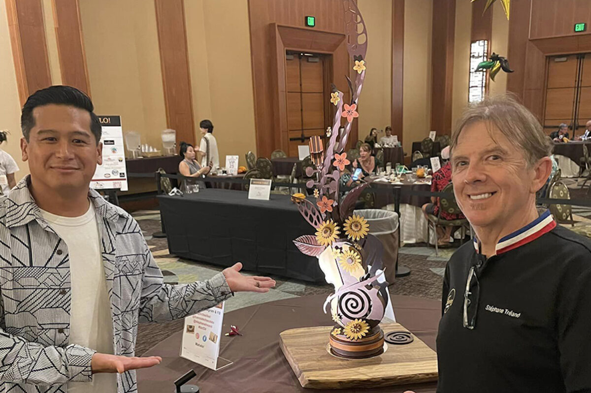 Two individuals stand on either side of an intricate chocolate sculpture on a table. The sculpture features various whimsical elements, including leaves and flowers. The room behind them has other tables and people, indicating a public event or gathering like one of the Kona Annual Events.