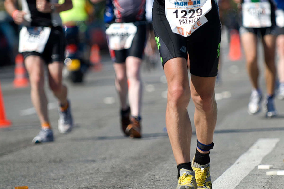 Runners in athletic gear participate in the Kona Annual Events long-distance race, passing orange cones on a paved road. One runner, wearing yellow shoes and a bib numbered 1229, is prominent in the foreground. The background features more runners in motion.