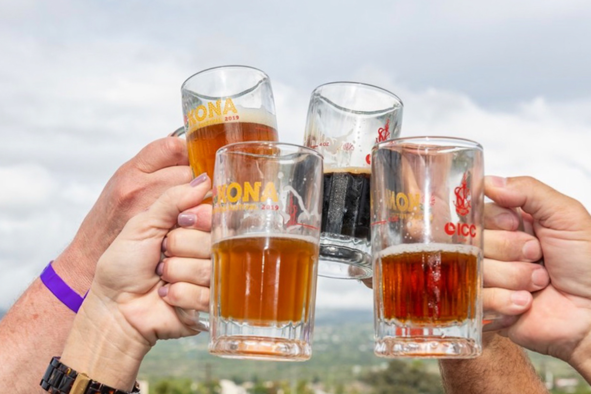 Four people clink their beer mugs together in a toast, with a scenic outdoor background. Each mug contains a different variety of beer, ranging from light to dark. The sky is cloudy, suggesting an overcast day at one of the Kona Annual Events.