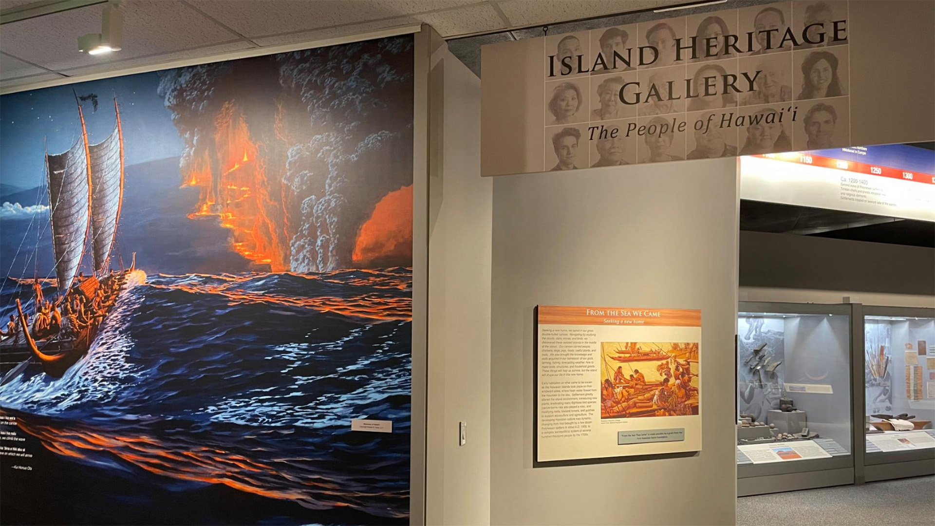 An exhibit in the Island Heritage Gallery titled "The People of Hawaiʻi." The foreground features a mural depicting a traditional Hawaiian canoe navigating rough seas near an erupting volcano. Nearby, there's an informative display with text and images.