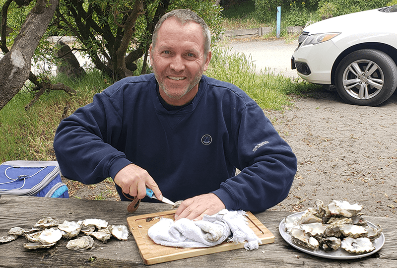A man smiles at the camera while shucking oysters at a picnic table. He wears a dark blue sweatshirt and holds an oyster knife in his hand. Several oysters rest on the table, and a car and greenery are visible in the background.