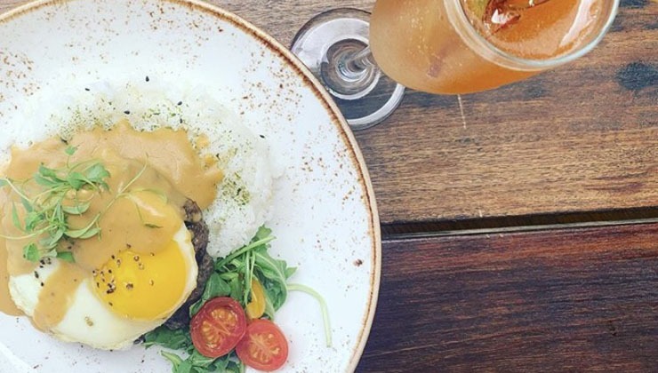 A wooden table holds a plate of gourmet food, featuring a sunny-side-up egg topped with a sauce, served over white rice, garnished with microgreens and cherry tomatoes. A stemmed glass with a caramel-colored drink is beside the plate.