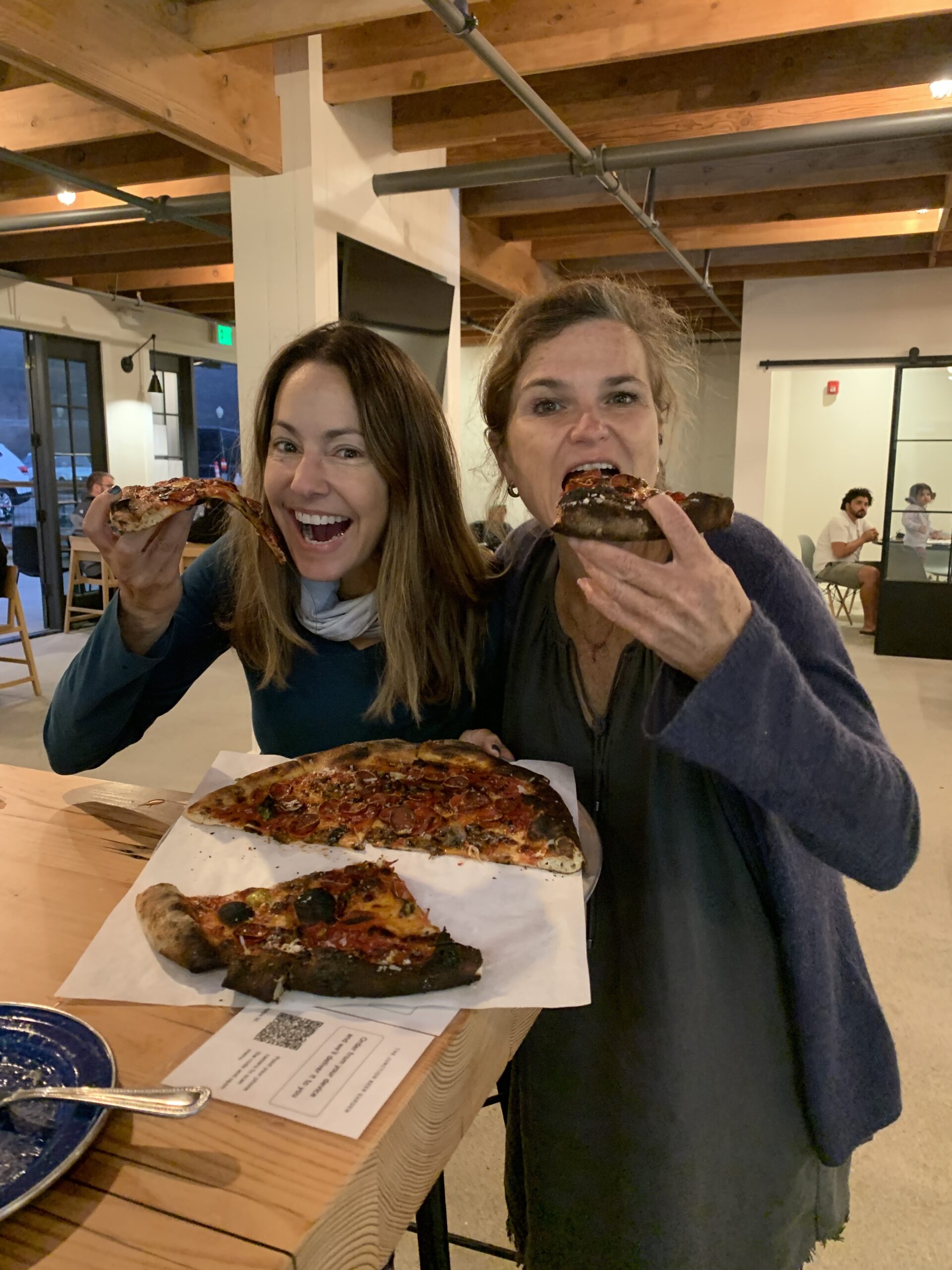 Two women smiling and holding slices of pizza in a casual indoor setting. They stand next to a wooden table with a partially eaten pizza resting on a white plate. The background features wooden beams and other diners.
