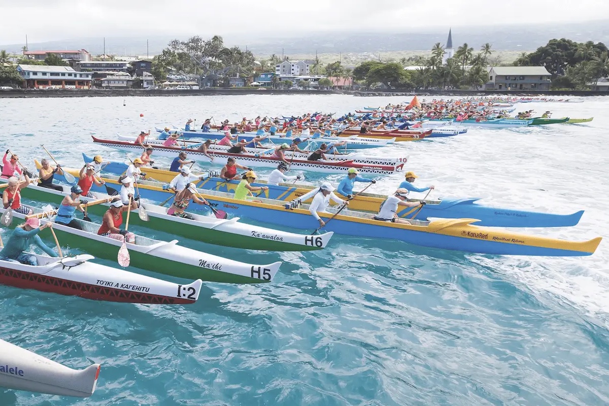 A vibrant canoe race with colorful canoes and paddlers wearing matching colored gear on a blue ocean. Multiple teams are competing in sync, with a coastal town and palm trees visible in the background under a clear sky, part of the exciting Kona Annual Events.