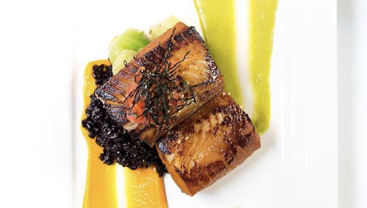 A gourmet dish featuring seared fish fillet on a bed of black rice and vegetables, garnished with herbs. The plate is artistically drizzled with vibrant yellow and green sauces. The presentation is minimalist and elegant.
