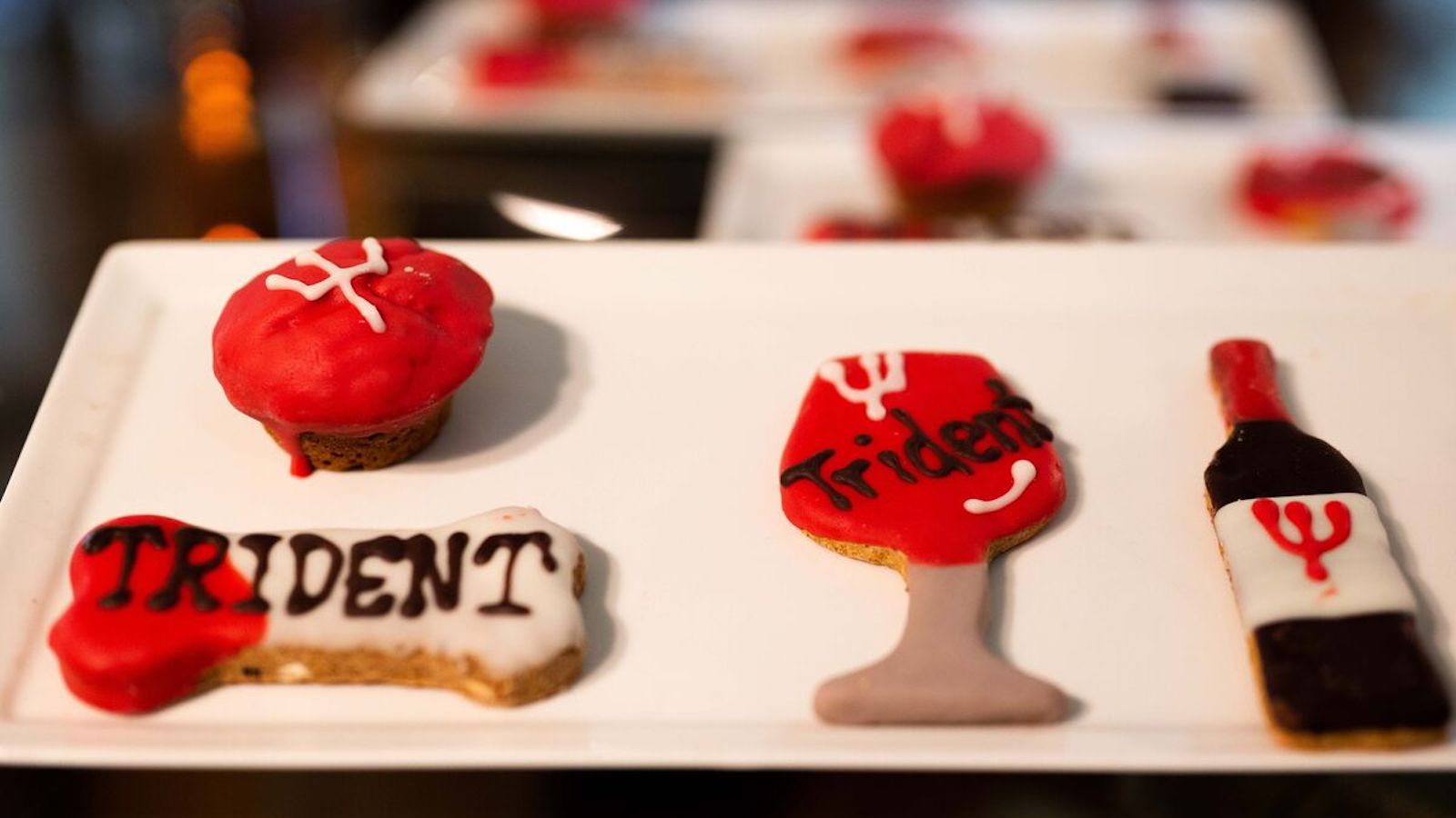 A rectangular white platter holds various cookies decorated with white, red, and black icing. Designs include a round cupcake, a bone-shaped cookie marked "TRIDENT," a wine glass-shaped cookie, and a bottle-shaped cookie featuring a red trident symbol—perfect for Napa Valley tasting rooms or dog-friendly gatherings.