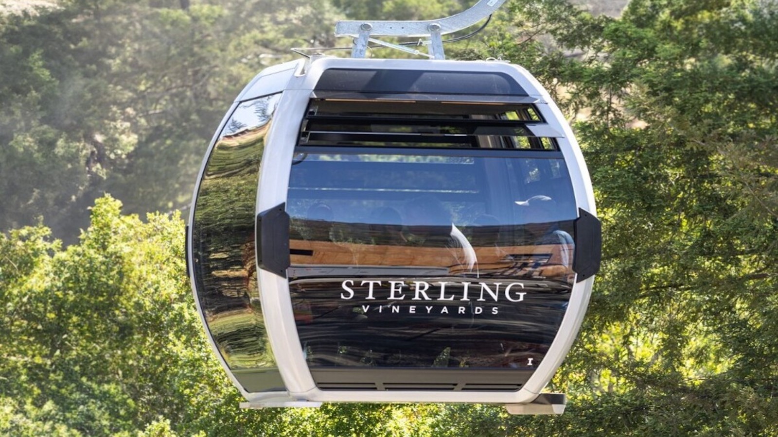 A modern gondola lift with "Sterling Vineyards" written on the side glides through lush green trees under sunny skies in Napa Valley, with reflections of the surrounding scenery visible on its glass windows. This dog-friendly experience offers a unique view of the renowned vineyard's tasting rooms.