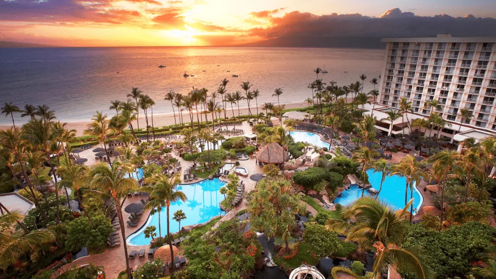 A tropical resort in Hawaii overlooking the ocean at sunset. The scene includes multiple swimming pools surrounded by palm trees and lush greenery. A large hotel building sits to the right, and the beach stretches out in the background with boats on the water, making it one of the best hotels with pools.