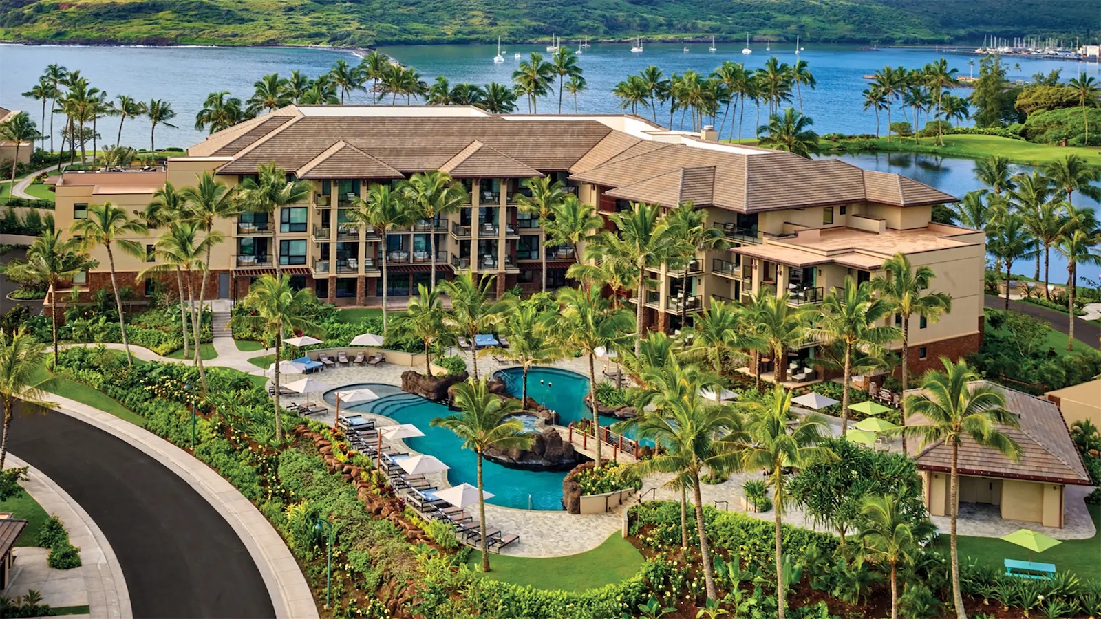 A luxury getaway in Hawaii, this resort is surrounded by lush greenery with a large swimming pool and sun loungers, overlooking a calm blue lake with sailboats. The tropical-style buildings are adorned with palm trees and mountains in the background under a clear sky.