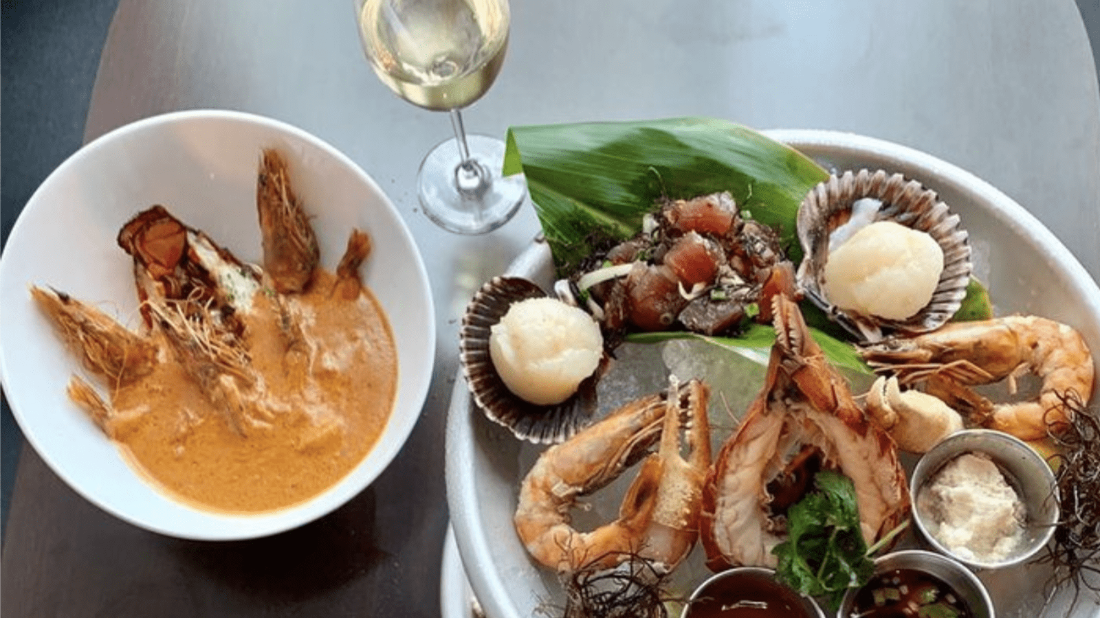 A seafood meal featuring a bowl of lobster bisque with whole lobster tails and a platter with shrimp, scallops, and fish garnished with greens and sauces, is served alongside a glass of white wine on a dark table.