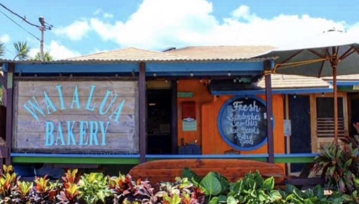 A small bakery with a rustic wooden exterior and a large sign that reads "Waialua Bakery". Another chalkboard sign advertises fresh sandwiches, smoothies, ice cream, and baked goods. Lush green plants are in the foreground, and a bright, sunny sky is overhead.