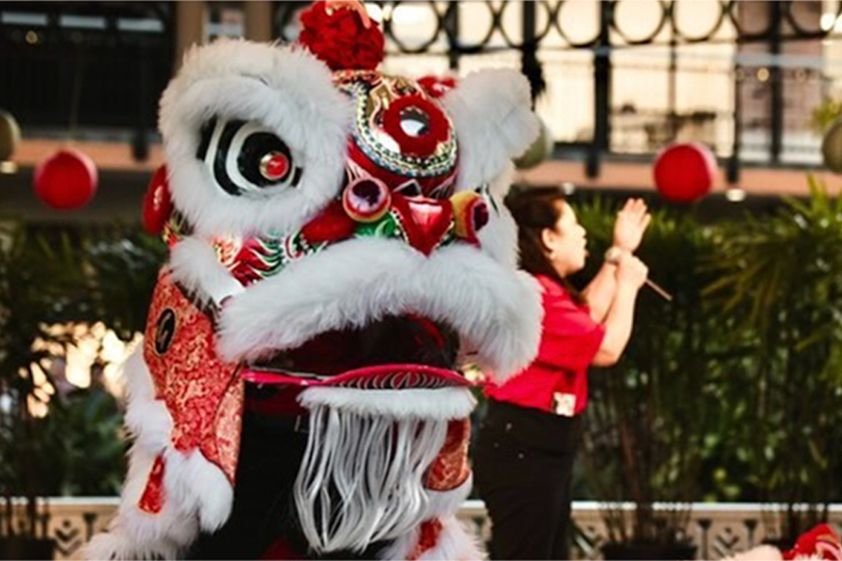A vibrant Chinese lion dance costume, with intricate red, white, and gold details and large expressive eyes, is being worn by performers at the Waikoloa Annual Events. In the background, a person in a red shirt is speaking into a microphone. Red lanterns and foliage are also visible.