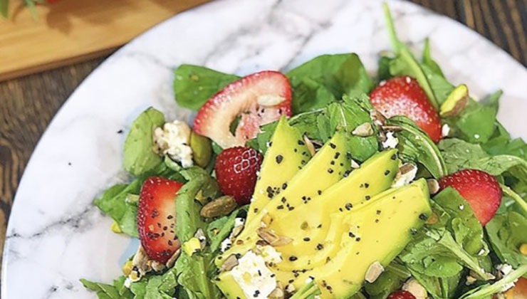 A close-up of a fresh salad featuring sliced avocado, strawberries, leafy greens, crumbled feta cheese, and sprinkled seeds on a white marble plate. The vibrant colors create an appetizing presentation.