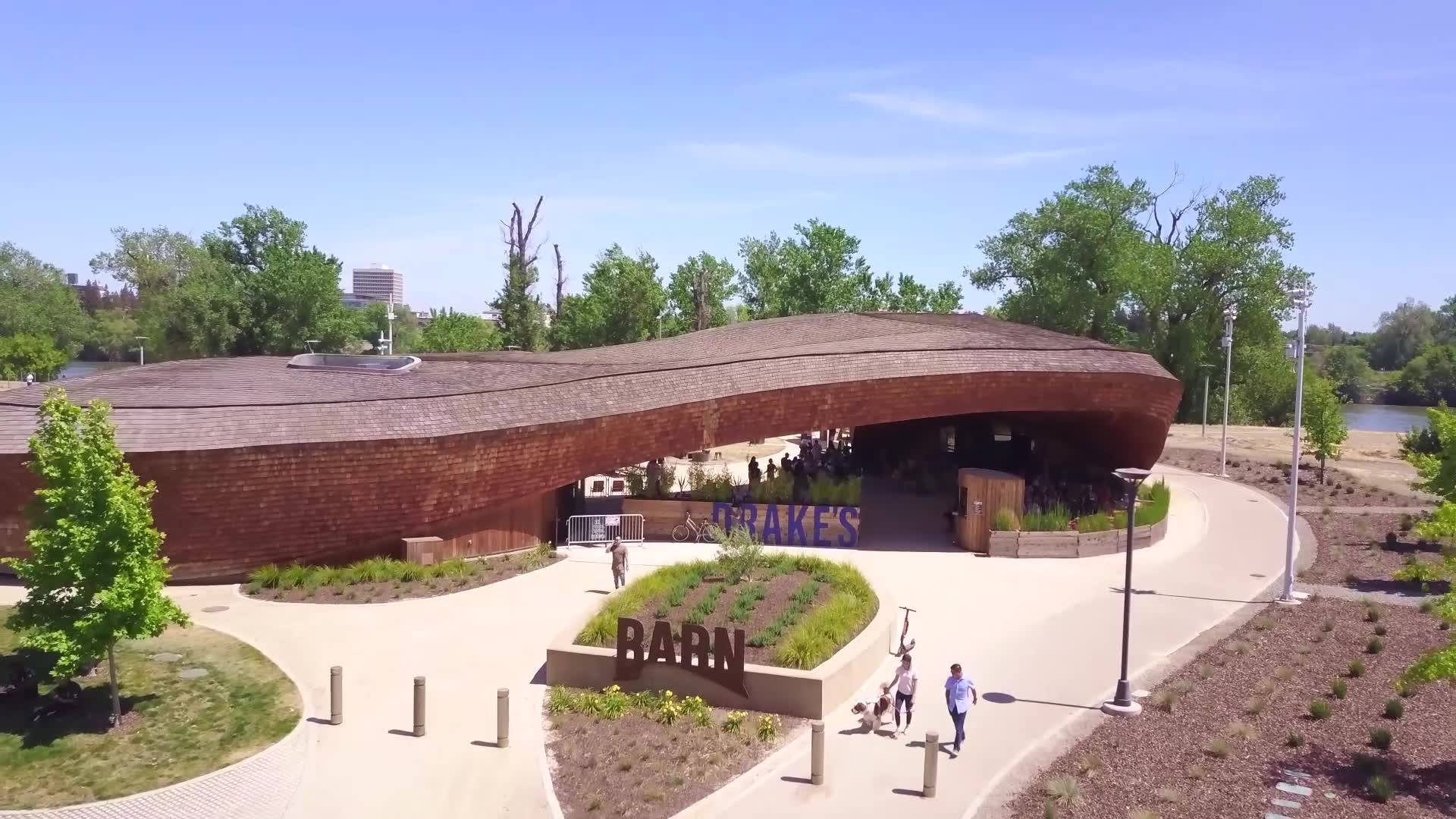 A large, modern wooden building with a curved roof labelled "Blake's Barn" sits amidst landscaped greenery on a sunny day. Several people walk on the pathways surrounding the building, which is famed for offering the best dessert in Sacramento. Trees are visible in the background.