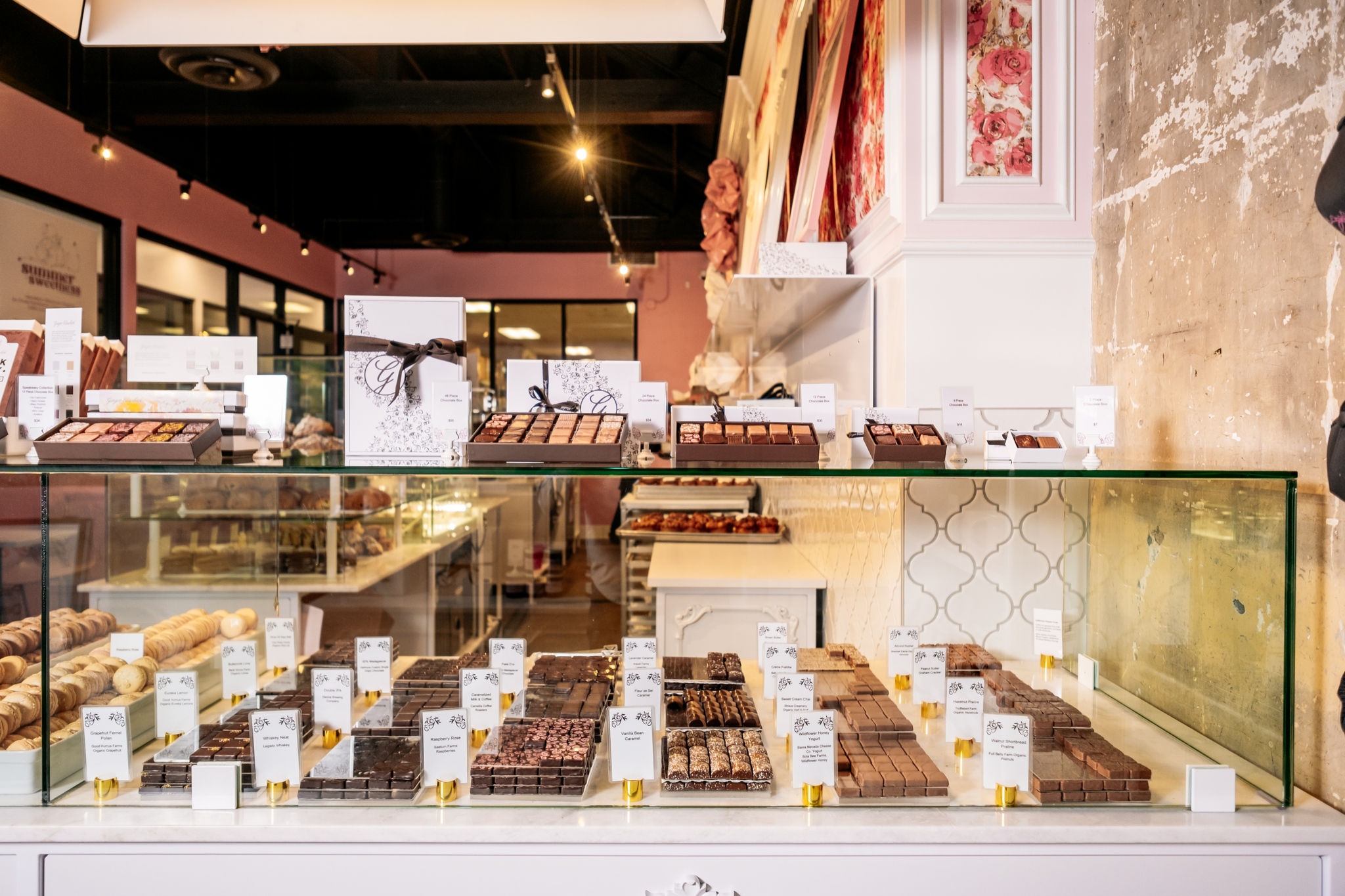 A glass display case in a bakery showcases an assortment of chocolates and pastries with labels. The background features more baked goods, a pink wall with floral decorations, and modern lighting, creating an inviting atmosphere perfect for those seeking the best dinner in Sacramento.