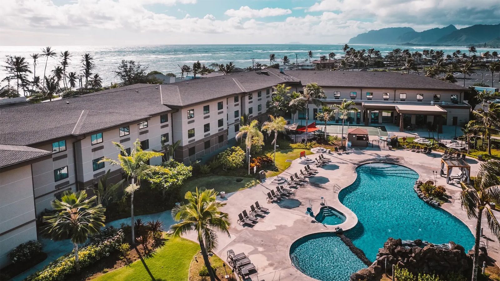 Aerial view of a seaside resort featuring a large, curvy outdoor pool surrounded by lounge chairs and palm trees. The multi-story hotel building overlooks the ocean, which is bordered by lush hills and mountains in the distance under a partly cloudy sky.