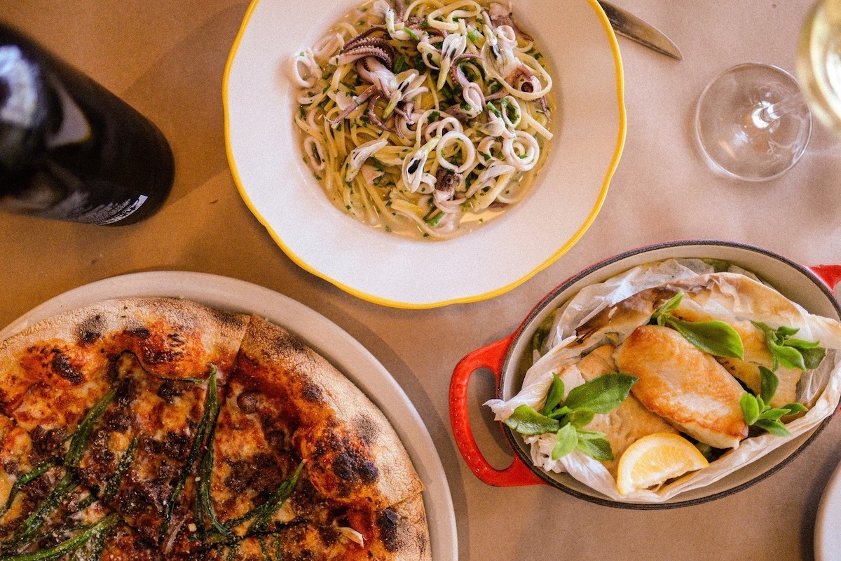 A table features three dishes: a pizza topped with green vegetables and cheese, a bowl of squid pasta in a white sauce, and a baked fish fillet garnished with fresh basil and served with a lemon wedge. A bottle of wine from Napa Michelin Restaurants and a glass of white wine are also visible.