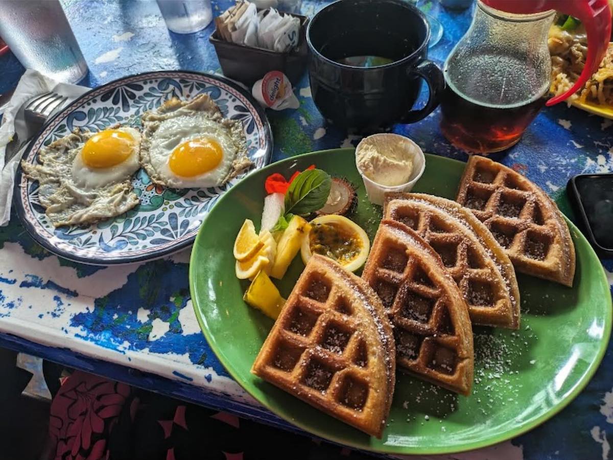 A plate of two sunny-side-up eggs sits on the left, next to a plate of four waffle quarters dusted with powdered sugar on the right. The waffles are accompanied by lemon slices, serving of butter, and a garnish. A pitcher of syrup and a mug are in the background.