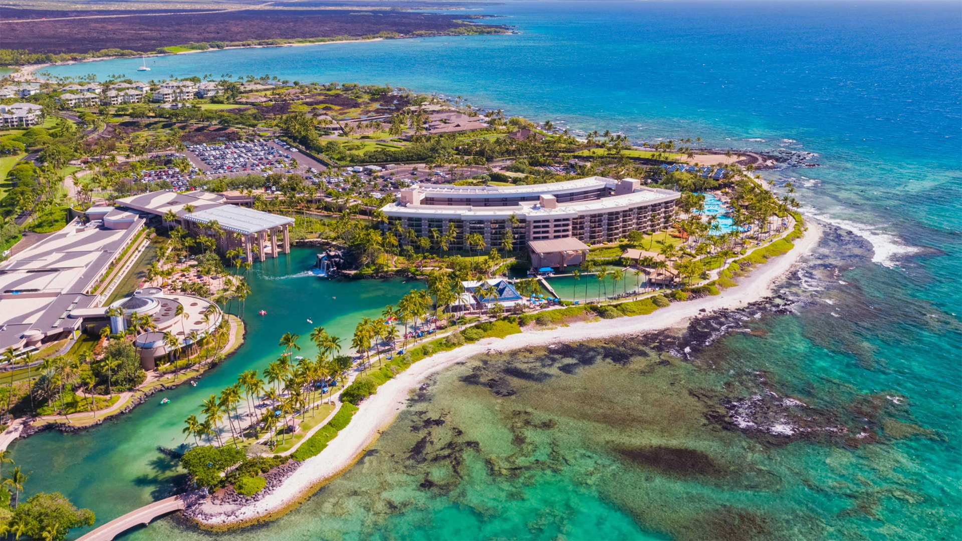 Aerial view of a tropical resort by the ocean, featuring a large, modern building surrounded by lush greenery and clear blue waters. The resort includes multiple pools, sandy beaches, and manicured gardens. Coral reefs and various facilities are visible in the vivid scene.