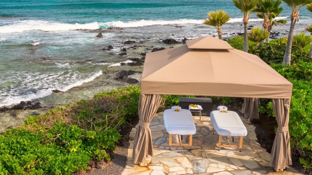 A beachside gazebo with two massage tables covered with white sheets, surrounded by lush greenery. The gazebo is set on a stone platform overlooking the ocean, with waves crashing on the rocky shoreline and palm trees in the background—perfect for those seeking Hotels with Spas in Hawaii.