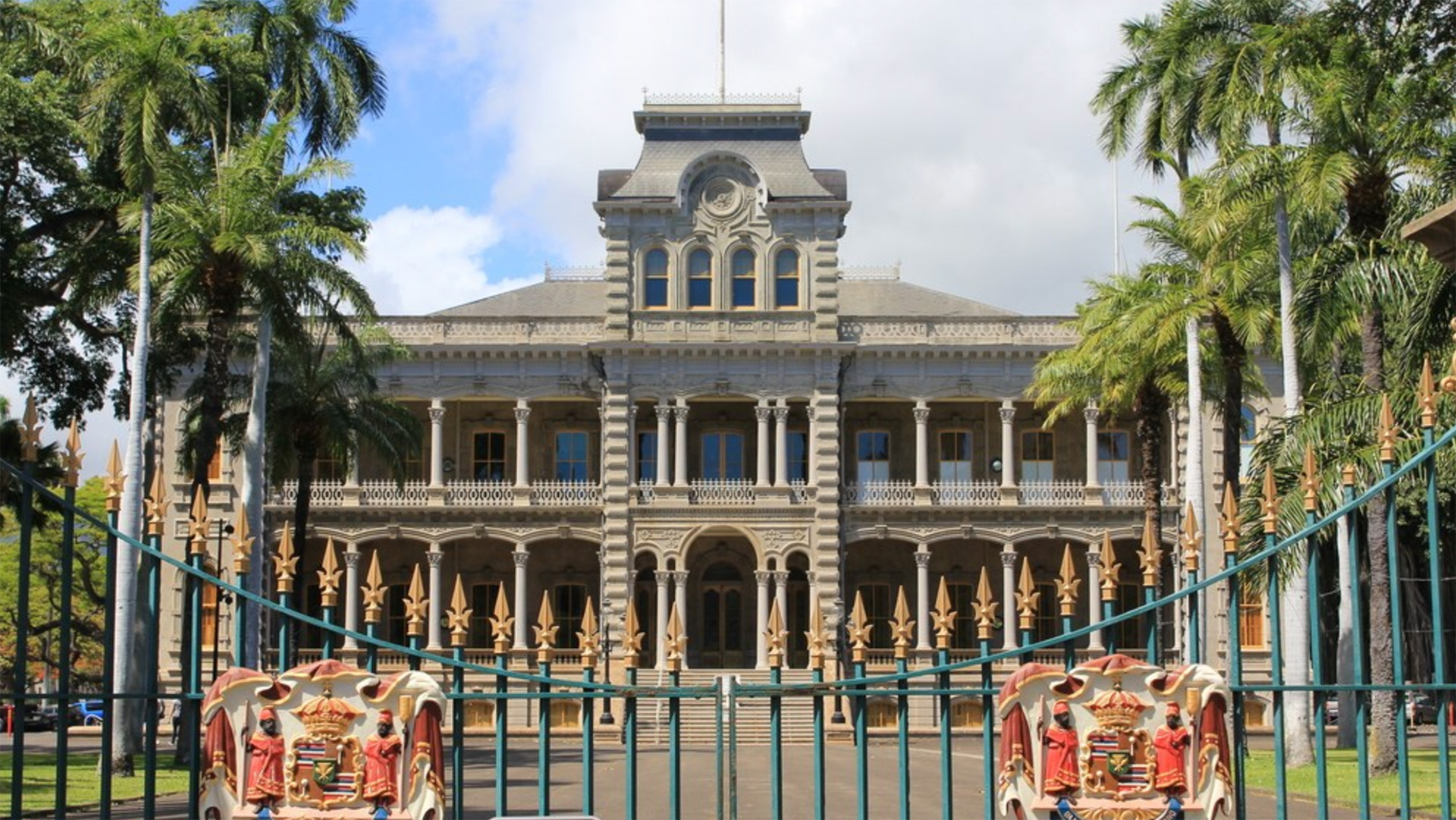 A grand historic building with ornate architectural details, Iolani Palace, framed by tall palm trees. An iron gate with the Hawaiian coat of arms in red and gold is partly visible in the foreground. The sky is partly cloudy.
