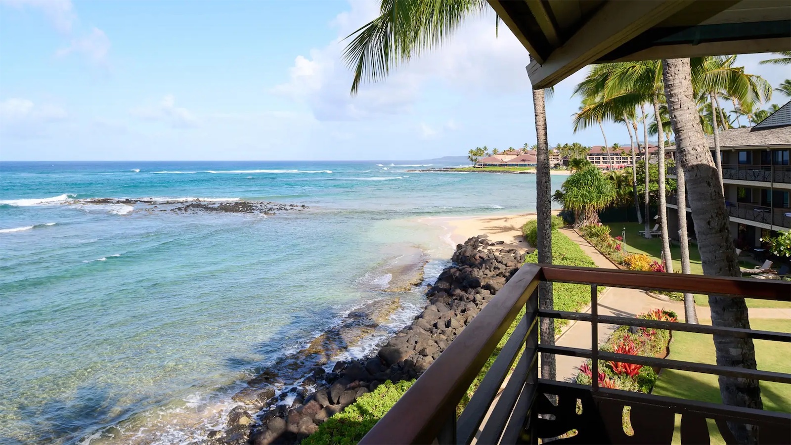 Image of a tropical oceanside view from a balcony. Clear blue water, white sandy beach, and palm trees line the shore. Several hotels with spas are visible in the distance along the Hawaiian coastline. The sky is clear with a few scattered clouds.