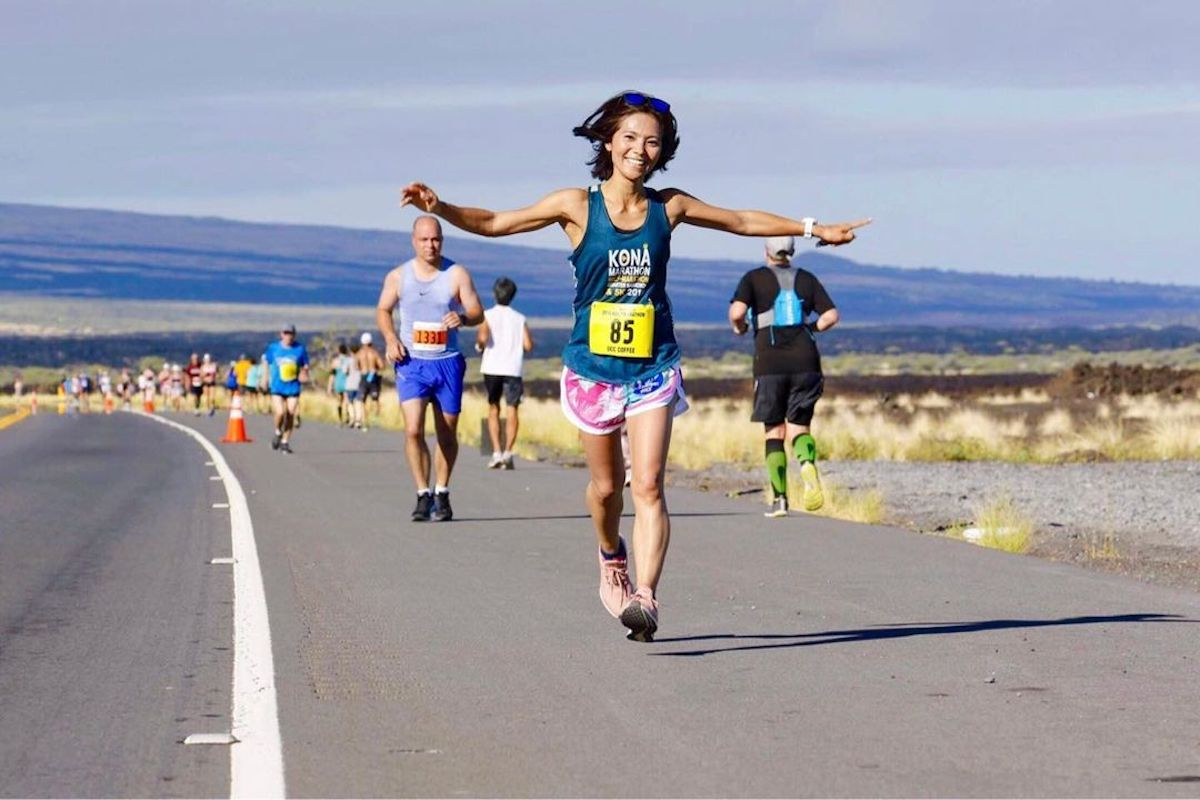 A woman in a blue tank top, pink and white shorts, and sunglasses runs joyfully with her arms outstretched on the highway during the Kona Annual Events race. Other runners are visible in the background. The surrounding landscape is dry and open, with distant hills under a clear sky.