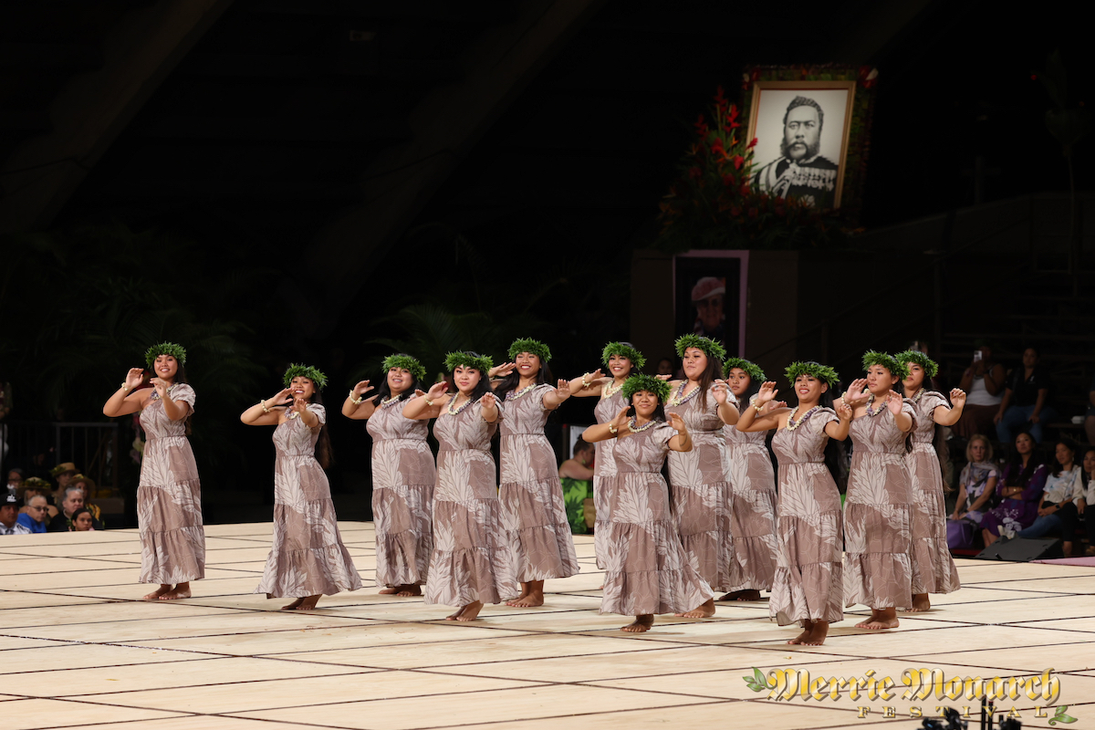 At the Hillo Annual Events, a group of dancers dressed in matching traditional Hawaiian attire, wearing long dresses and head leis, perform a synchronized hula dance on a stage with a large portrait and floral arrangement in the background. An audience watches from the sidelines.