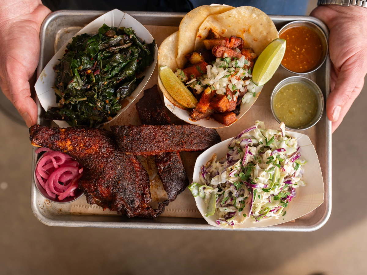 A person holds a tray of assorted food items, reminiscent of Napa's renowned Michelin-rated restaurants, featuring seasoned meat, pickled onions, sautéed greens, two tacos with toppings, and a side of coleslaw. Small containers of green and orange sauces are also on the tray.