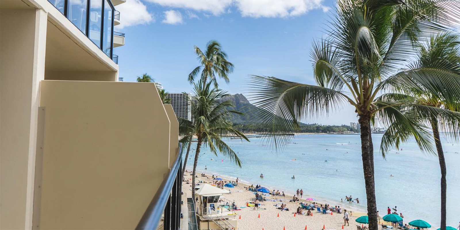 View from a balcony overlooking a tropical beach with clear blue waters. Palm trees sway in the breeze, and people are relaxing on the sand and swimming. A mountainous island landscape is visible in the distance under a bright, partly cloudy sky.