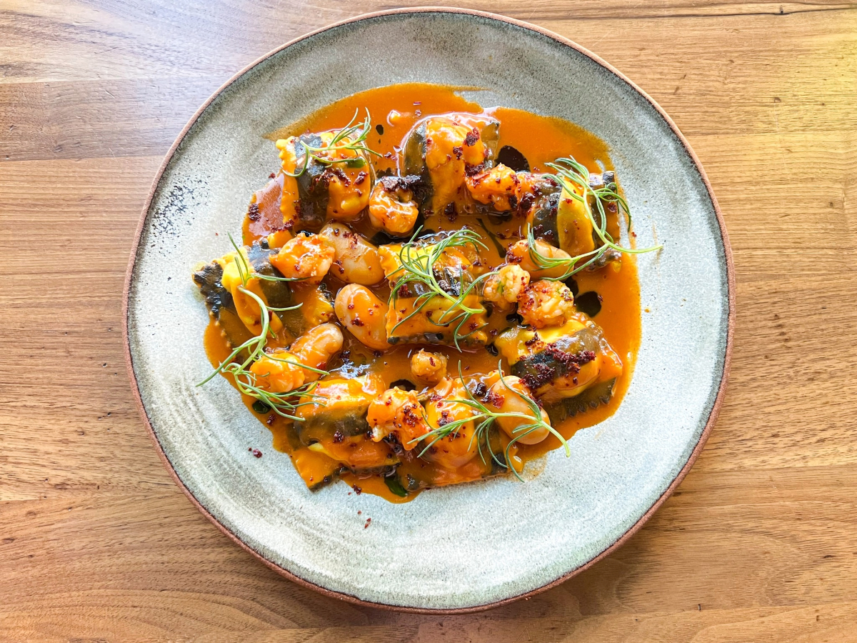 A plate of grilled seafood, possibly shrimp and scallops, garnished with thin green herbs and drizzle of rich, amber-colored sauce. The dish evokes the coastal charm of Half Moon Bay and is served on a simple, rustic plate set on a wooden table.