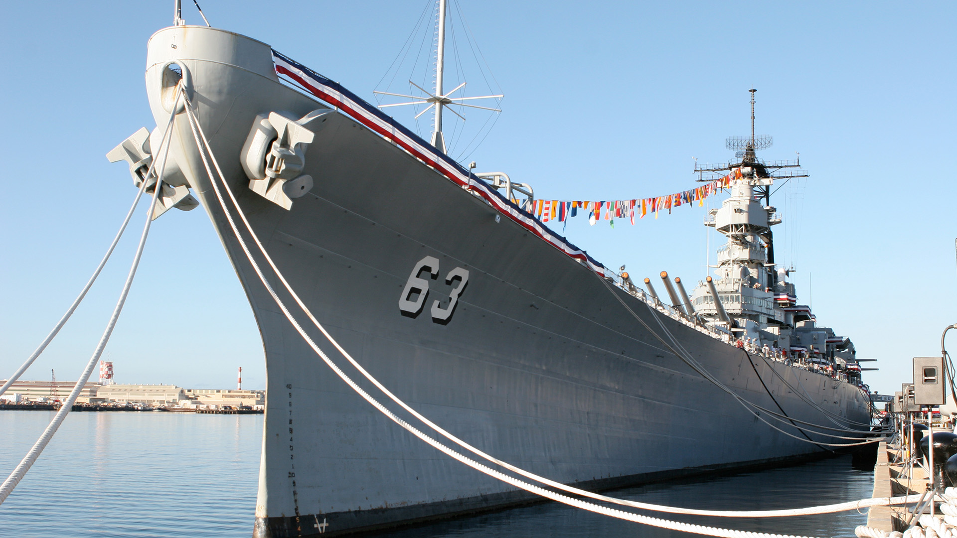An imposing gray naval battleship, marked with the number "63," docked at a harbor. The ship is adorned with decorative flags strung along its length. Multiple ropes secure it to the dock, and clear blue sky enhances the serene backdrop.