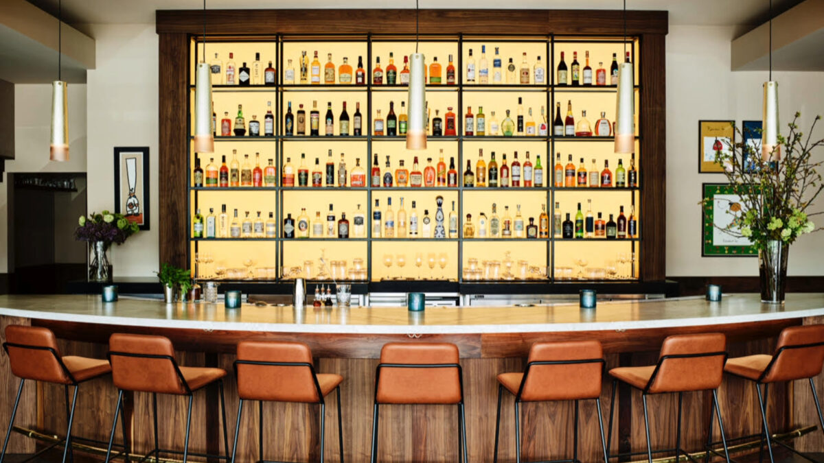 A stylish bar in Half Moon Bay features a well-lit, extensive collection of bottles on shelves behind a polished wooden counter. Seven padded brown stools line the counter, and modern pendant lights hang from the ceiling. Flower arrangements are at both ends of the counter.