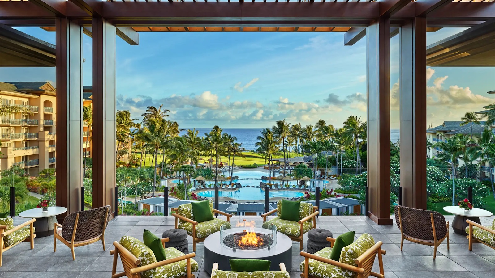 A luxurious outdoor lounge with wicker furniture and patterned cushions surrounds a fire pit, overlooking a pristine resort pool and tropical landscape leading to the ocean. Palm trees and well-maintained gardens enhance the serene and inviting atmosphere of this luxury getaway in Hawaii.