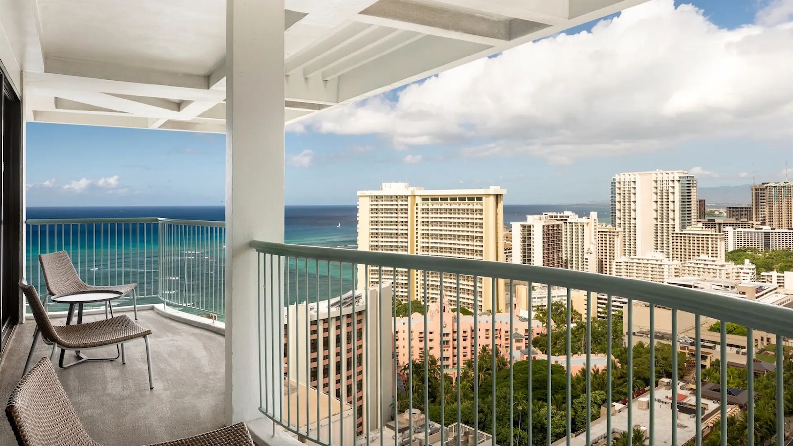 A spacious balcony with lounge chairs overlooks a city of high-rise buildings and offers a stunning ocean view. The sky, filled with scattered clouds, enhances the serene and picturesque scene, reminiscent of the tranquil beauty found in value hotels across the Hawaiian Islands.