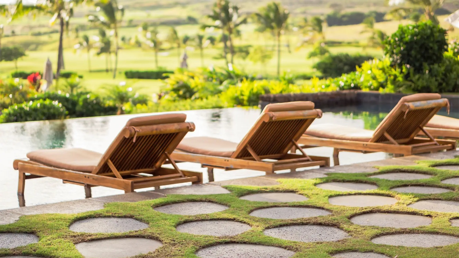 Three wooden lounge chairs with cushions are lined up by an infinity pool overlooking a lush, green landscape in Hawaii, complete with palm trees and a distant golf course. The foreground features a pathway with large circular stepping stones set in grass, reminiscent of luxurious hotels with spas.