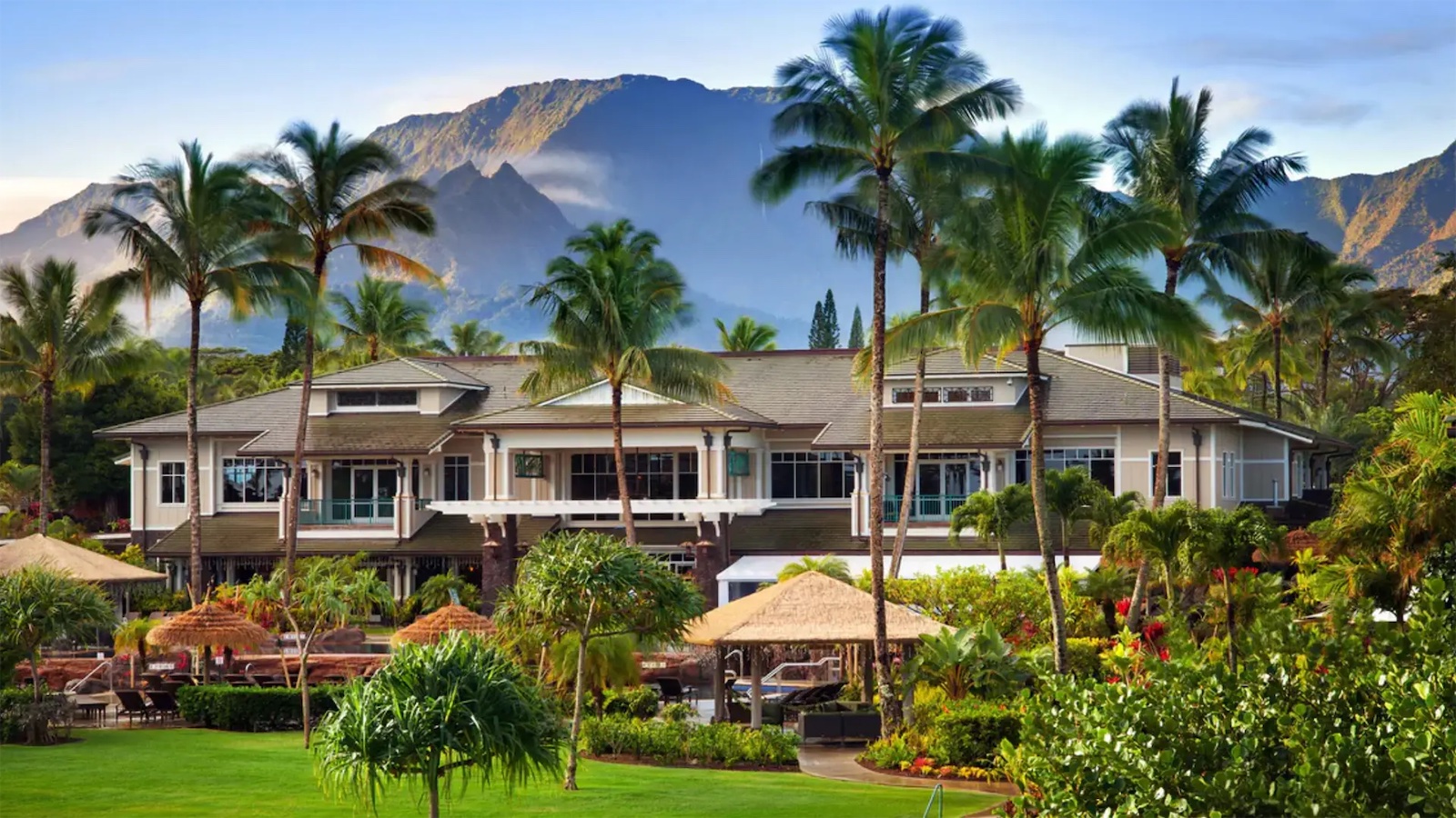 A luxurious tropical resort with a large, elegant building surrounded by palm trees and lush greenery. Thatch-roofed huts and well-maintained lawns are in the foreground. A mountain with a clear blue sky and scattered clouds is visible in the background.