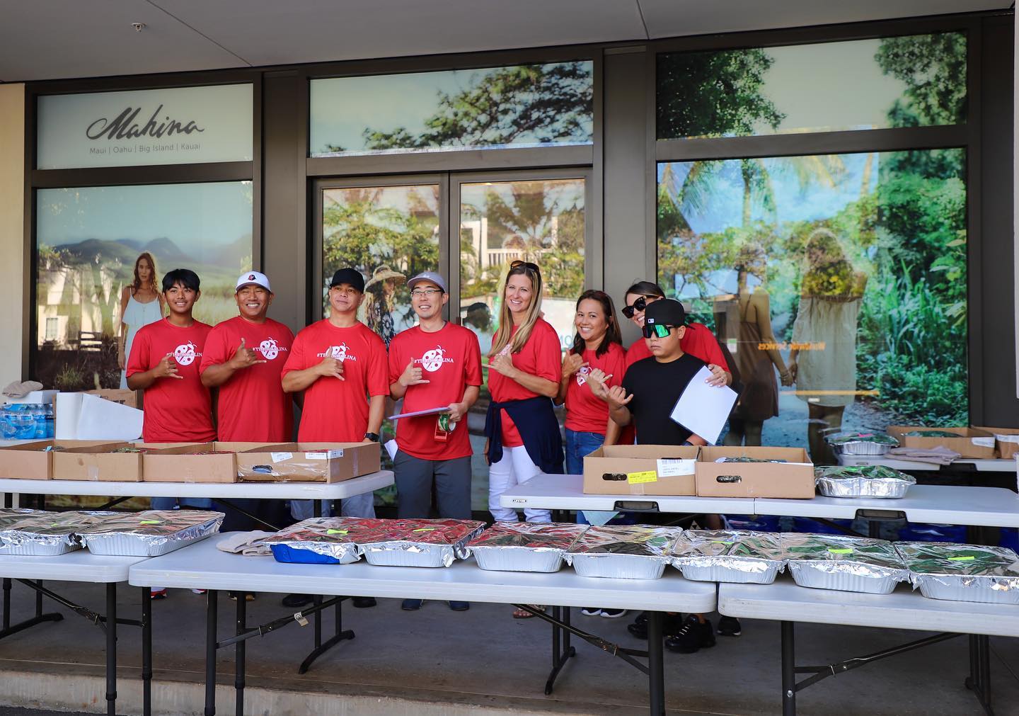 A group of volunteers wearing red T shirts stand in front of tables with prepared food for Annual Thanksgiving Outreach.
