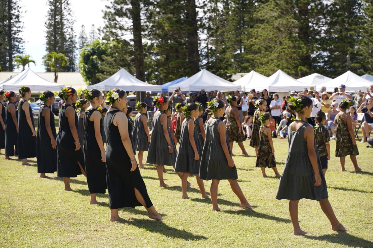 A large group of performers all wearing black stand on a lawn with booths with white tents in the background.