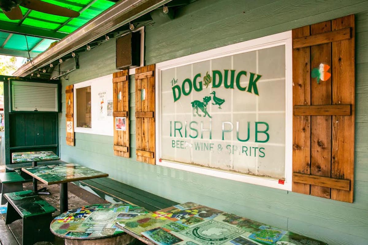 An outdoor patio for the Dog & Dog Irish Pub with a decal on the window that has their name and green tables and benches.