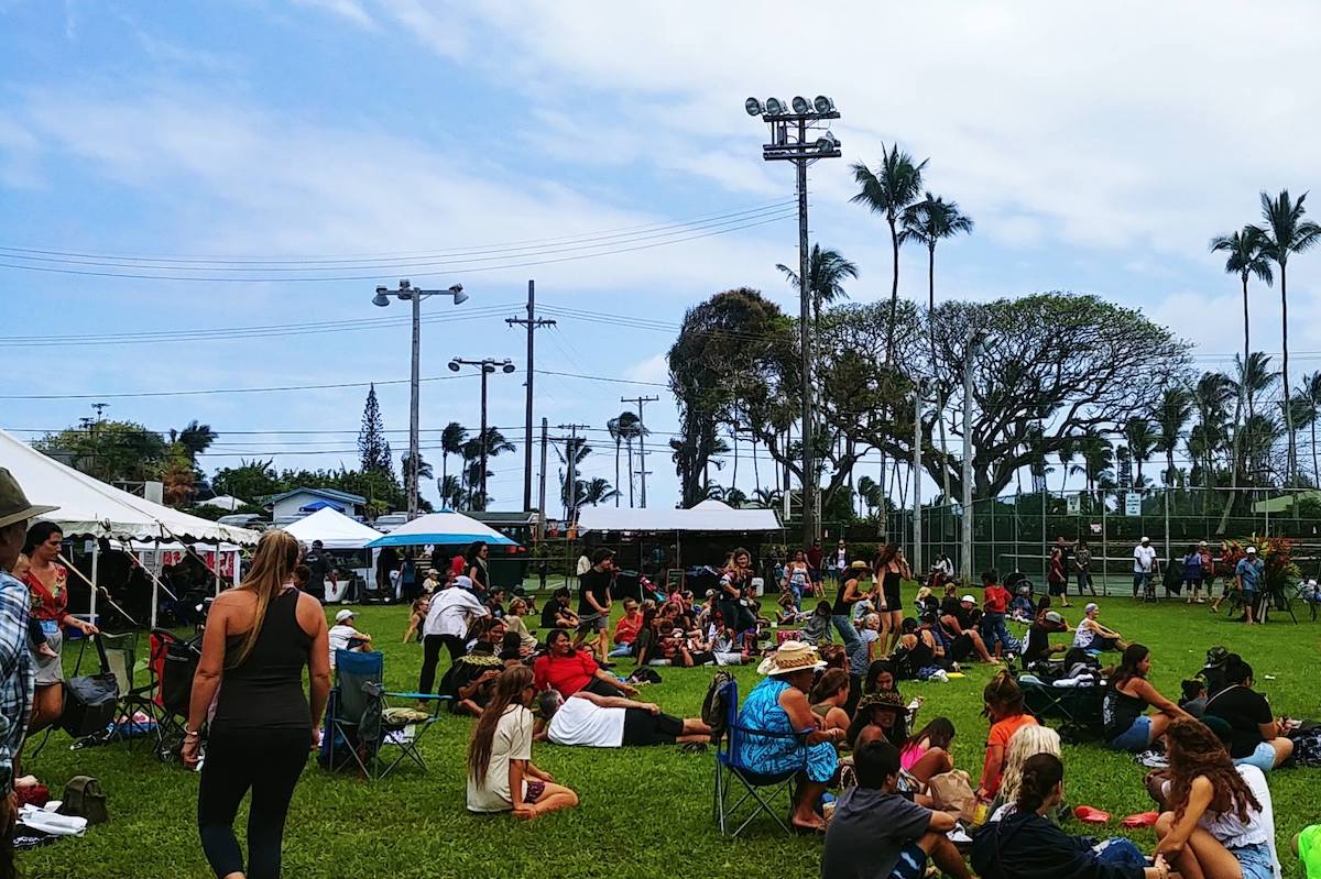 A lawn is filled with people walking around and sitting, with booths and palm trees in the background.