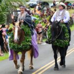 There are two people on horseback with leis riding through the streets of South Maui for the Festivals of Aloha, with a crowd watching.
