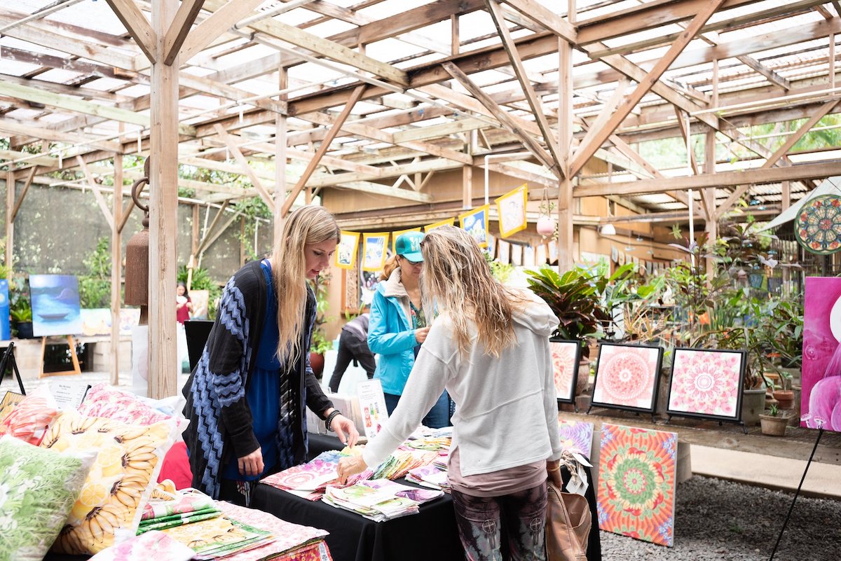 A vendor and visitor are at a booth for the Maui Open Studios event with lots of plants in the background.
