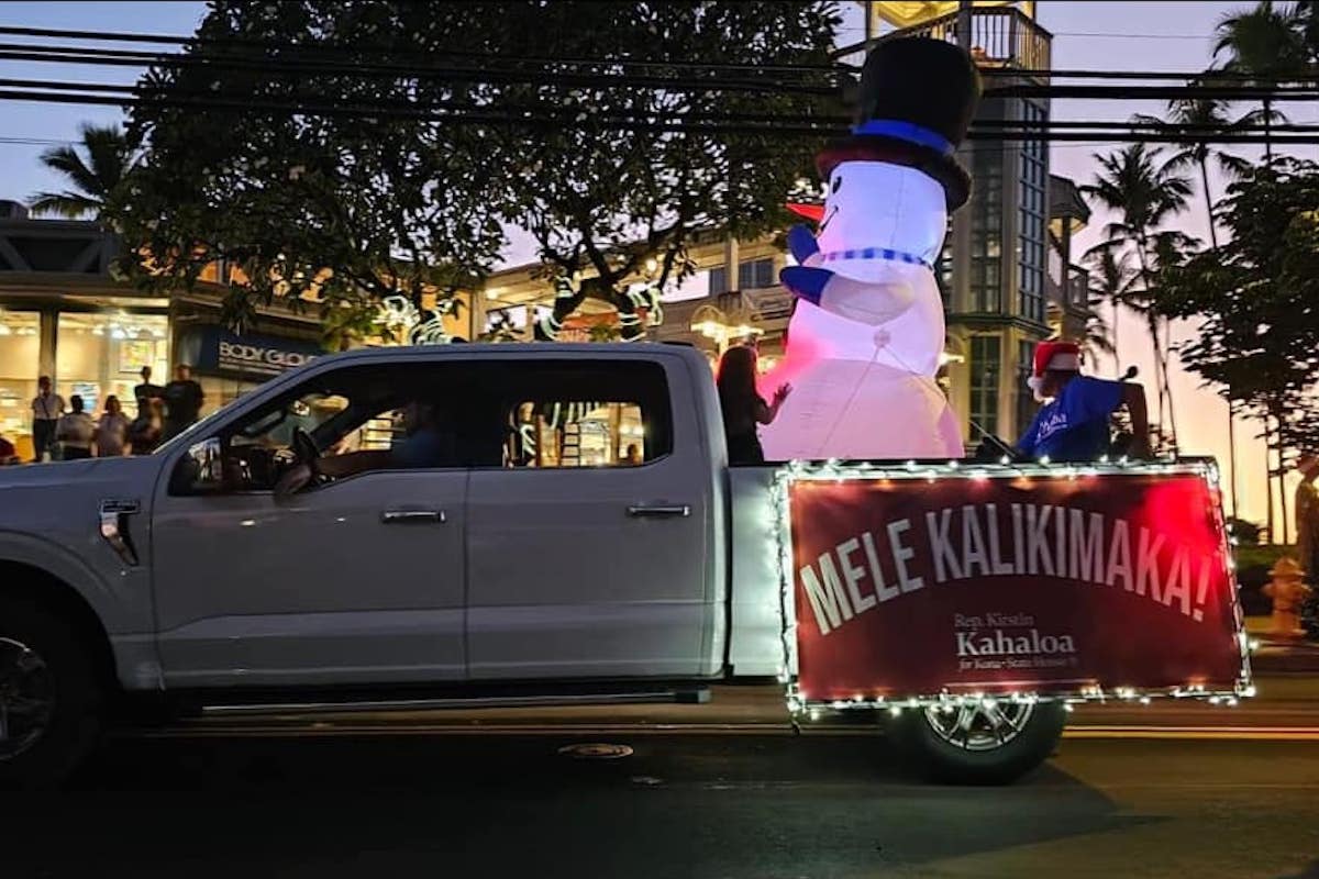 A white truck with a sign that reads "Mele Kalikimana" on the side of the truck bed with a lit and blown up snowman in the back.