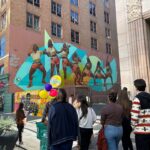 A group of people stand on a sidewalk and look up at a colorful mural on the side of a tall building.