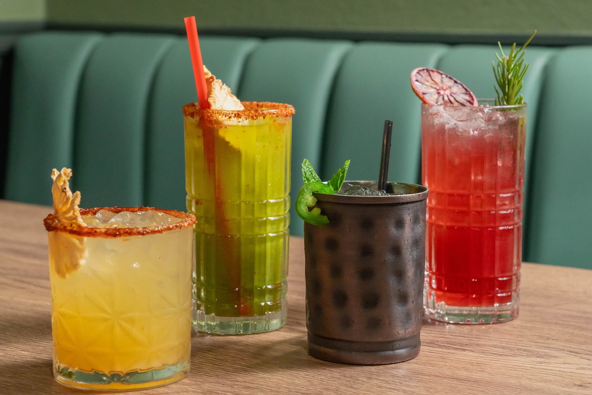 Four cocktails in various sized cups and of various colors including yellow, green, dark gray and red sit on a table with a green upholstered booth behind it.