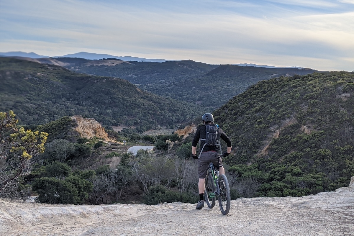 A man wearing a helmet sits on his bike on the edge of a cliff overlooking mountains.